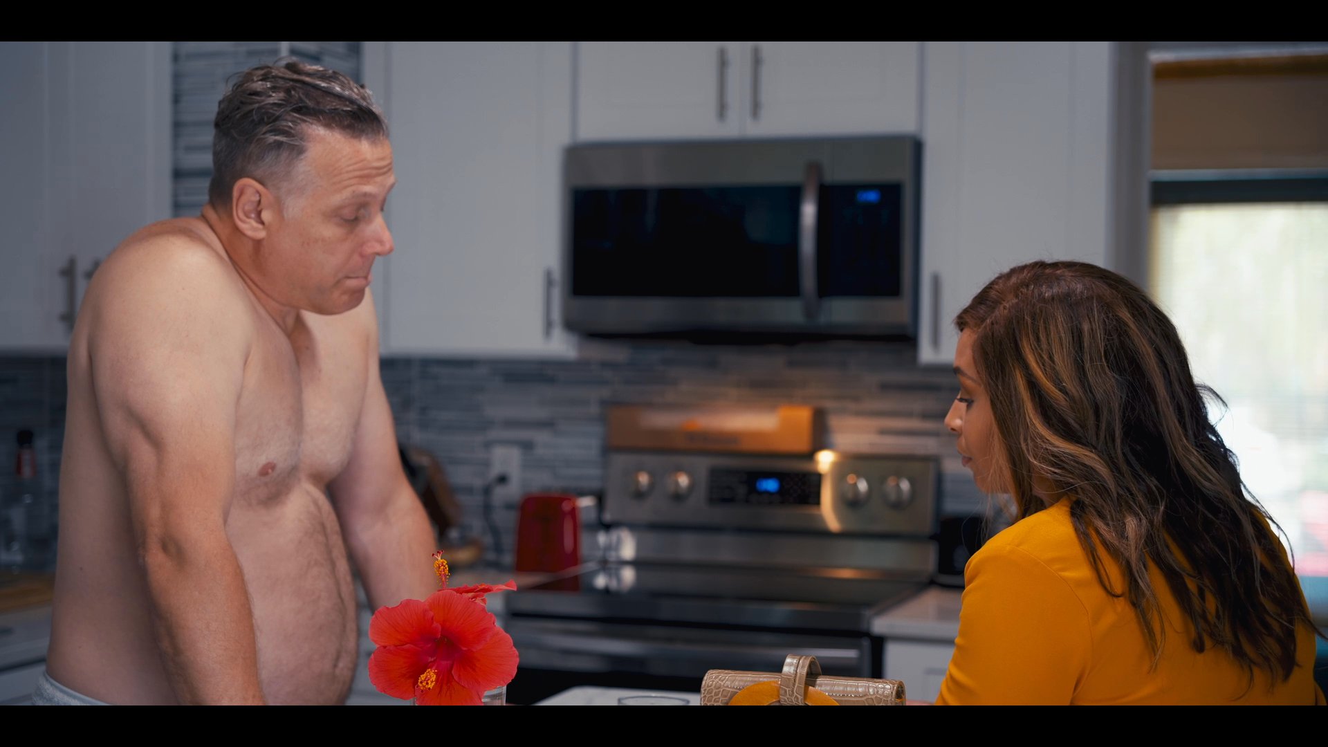 A man and a woman engage in a focused conversation while standing in a modern kitchen. The woman gestures while speaking as they maintain eye contact throughout the interaction.