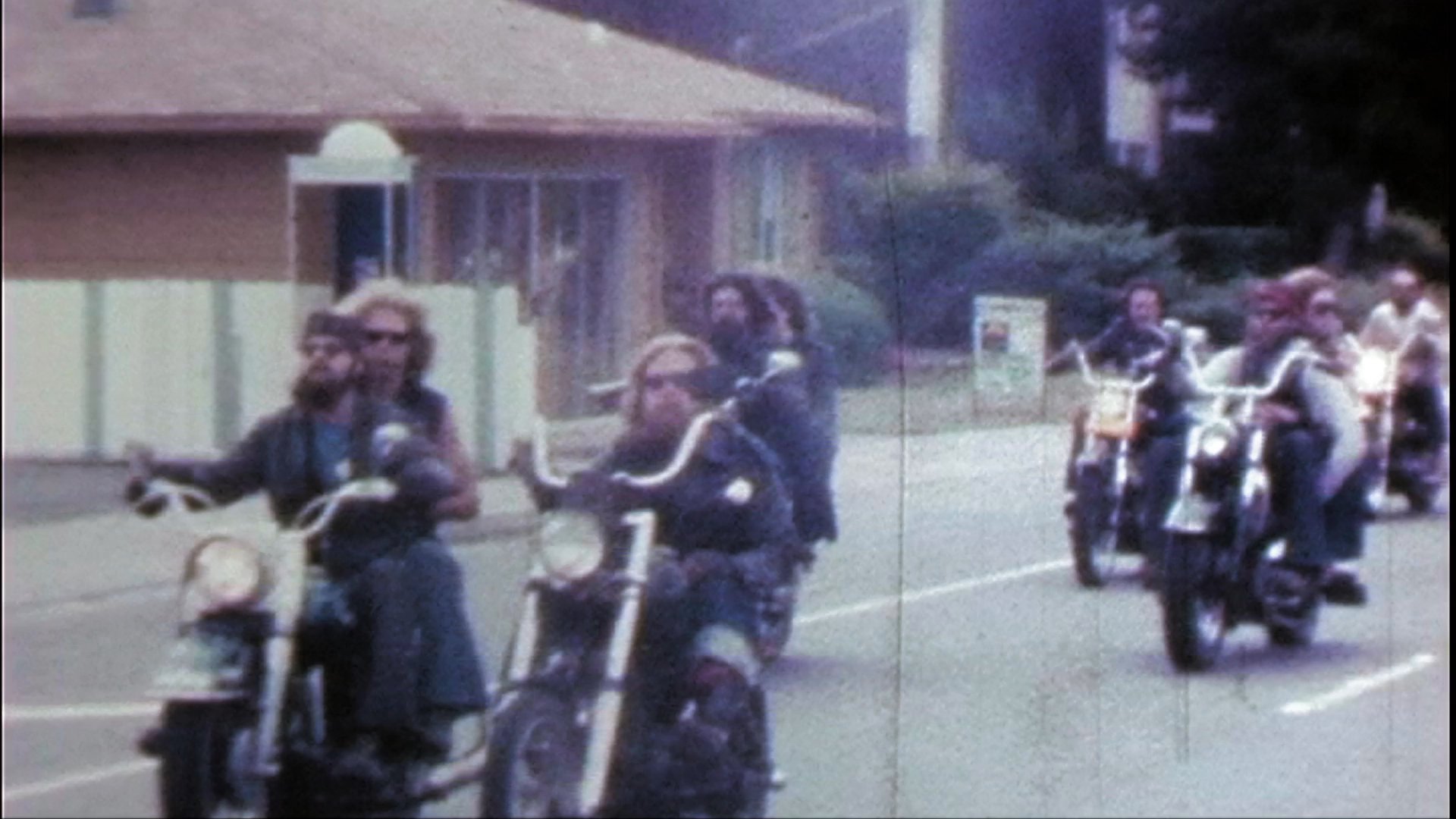 A group of motorcyclists riding custom choppers arrives at a commercial parking lot. The riders navigate their vintage motorcycles through the paved area in a coordinated formation.