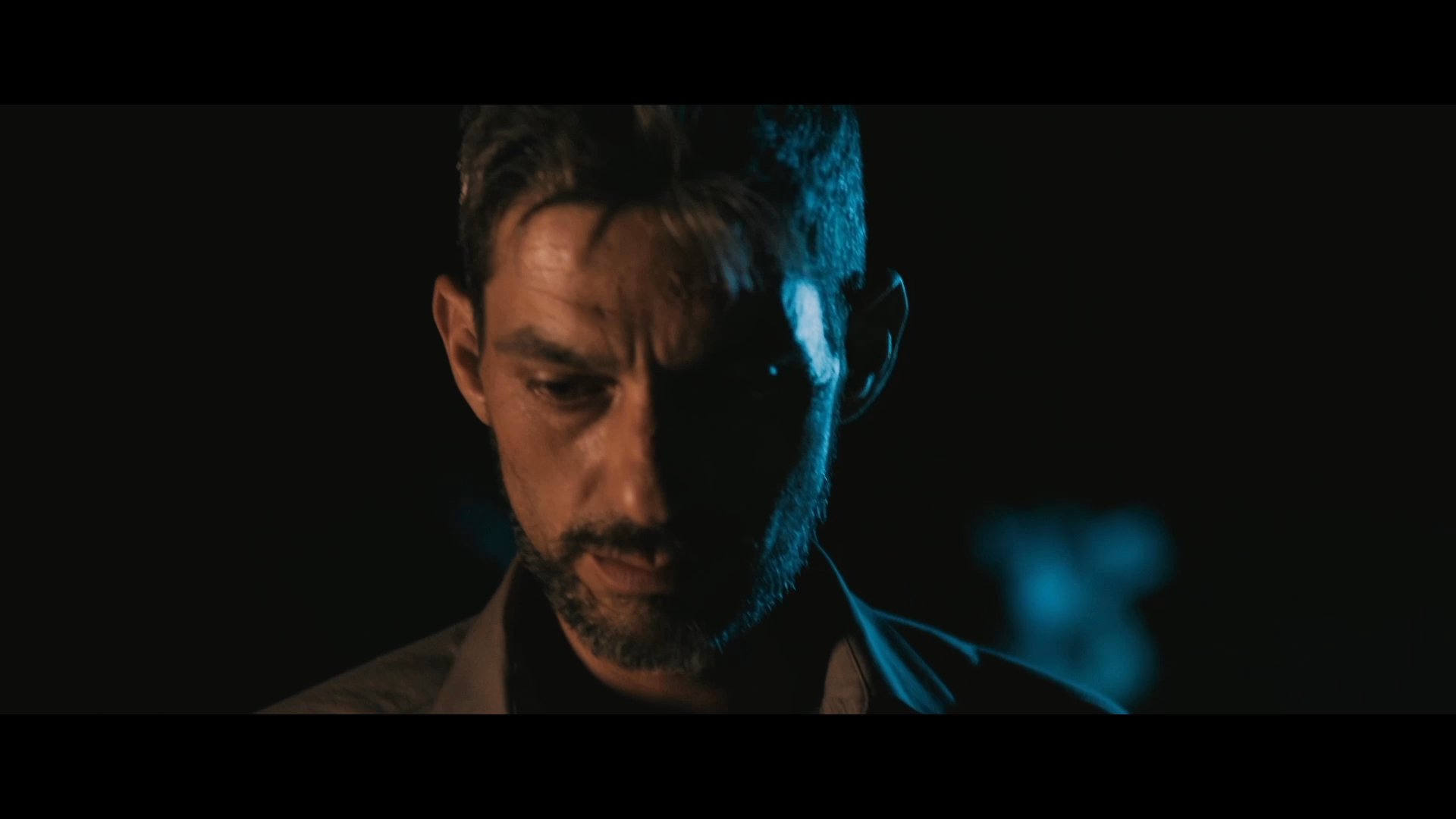 A man exhibits a pensive and somber expression in a low-light environment, contrasting with a woman sleeping peacefully in bed.