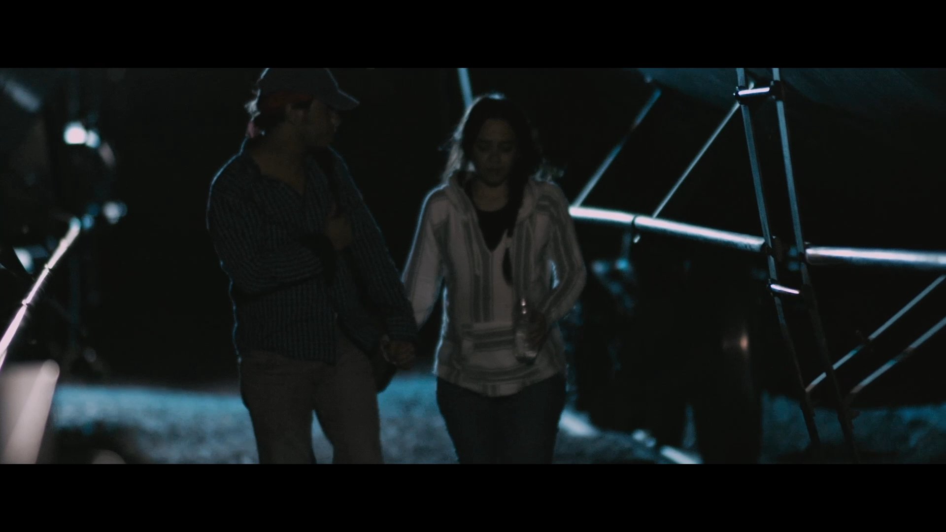 Two individuals walk along a railway track at night while discussing their safety and recent escape from a train. The scene highlights their tense interaction and relief after disembarking.