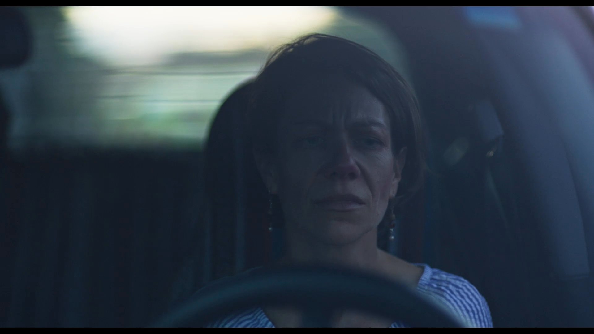 A woman receives a tender kiss on the cheek from a younger companion inside a vehicle before enduring a somber, solitary moment of reflection. The sequence captures an intimate farewell characterized by deep emotional weight and quiet intensity.