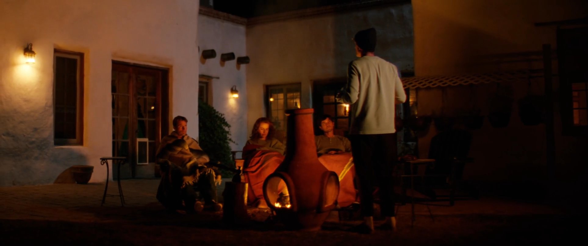 A man shares stories with a small group gathered around a warm fire in a desert courtyard at night. The intimate setting encourages reflection and shared experience among the companions.