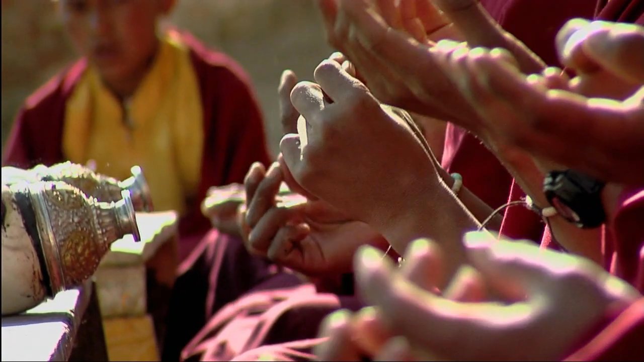 Young monks gather in a monastery courtyard to engage in traditional prayer and ritual activities, observed by an interviewer. The sequence transitions between the communal ceremony and a focused individual account of the cultural practice.