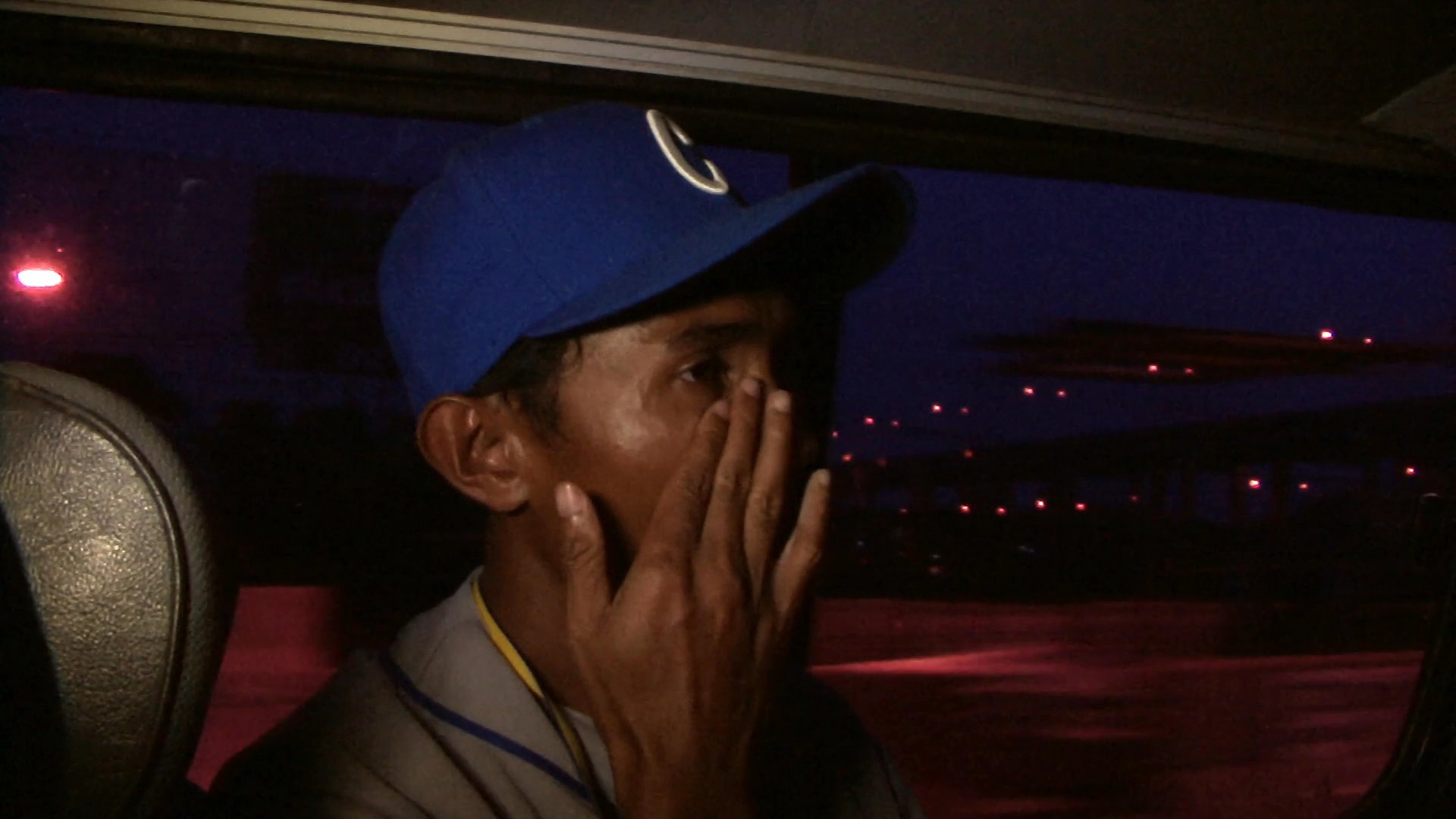 A man wearing a baseball cap sits in a vehicle at night, looking out the window while lost in thought during his commute.