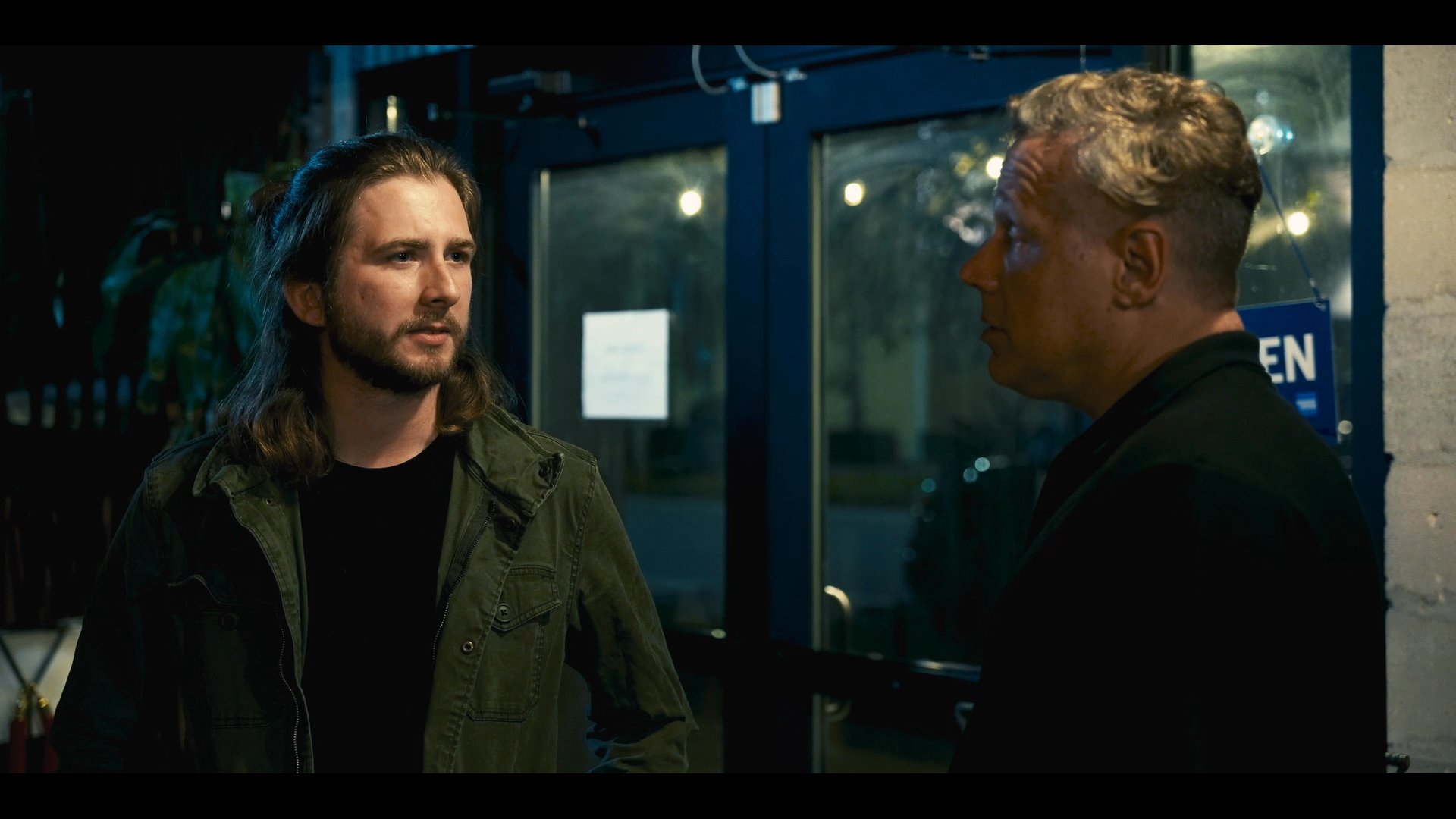 Two men engage in a serious conversation outside a commercial establishment at night, later joined by a third individual to continue the discussion.