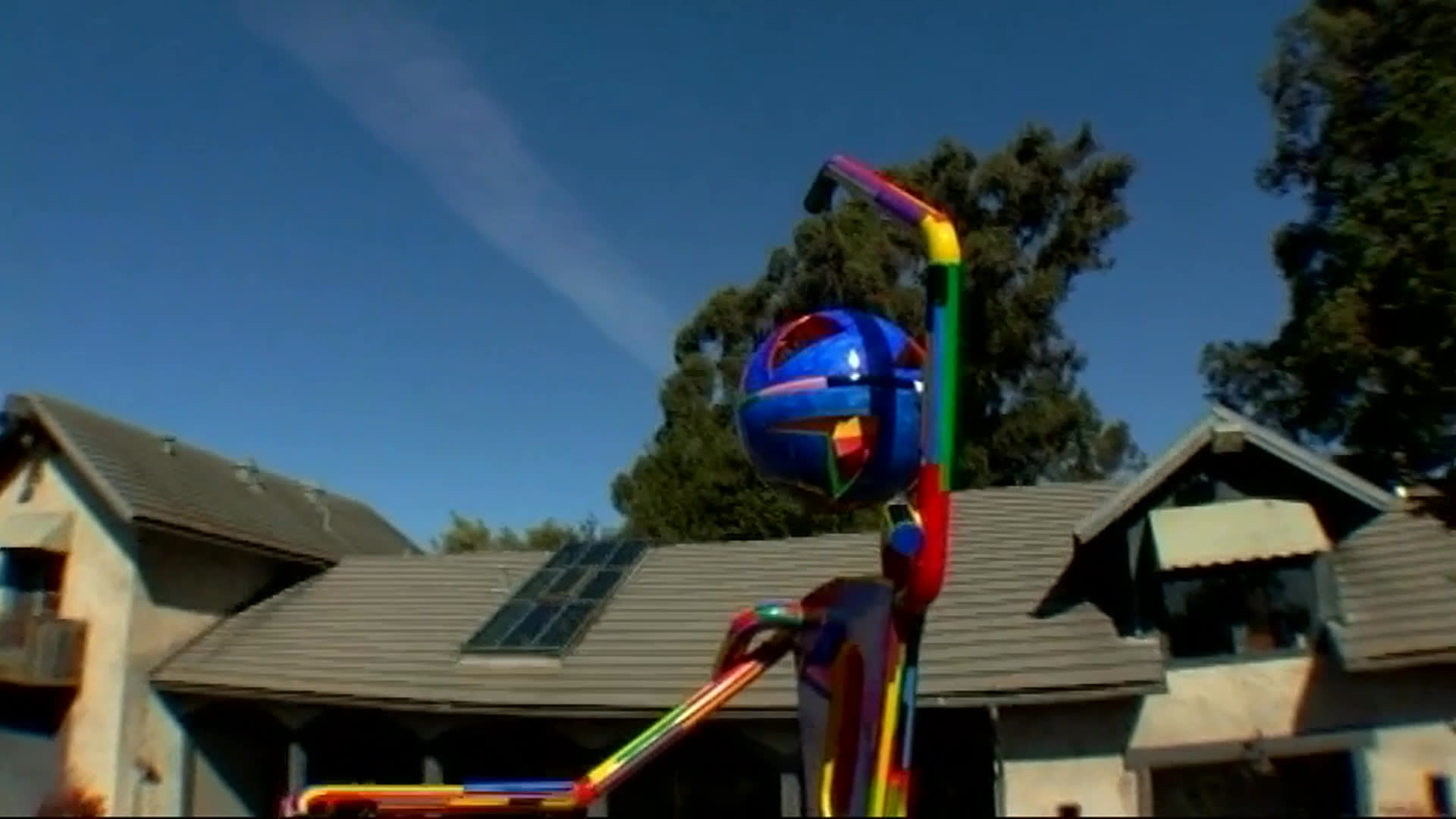 An older man discusses a brightly colored human-form sculpture displayed outside a house. Its hanging pieces shift gently in the wind as the view moves from the full installation to its details.
