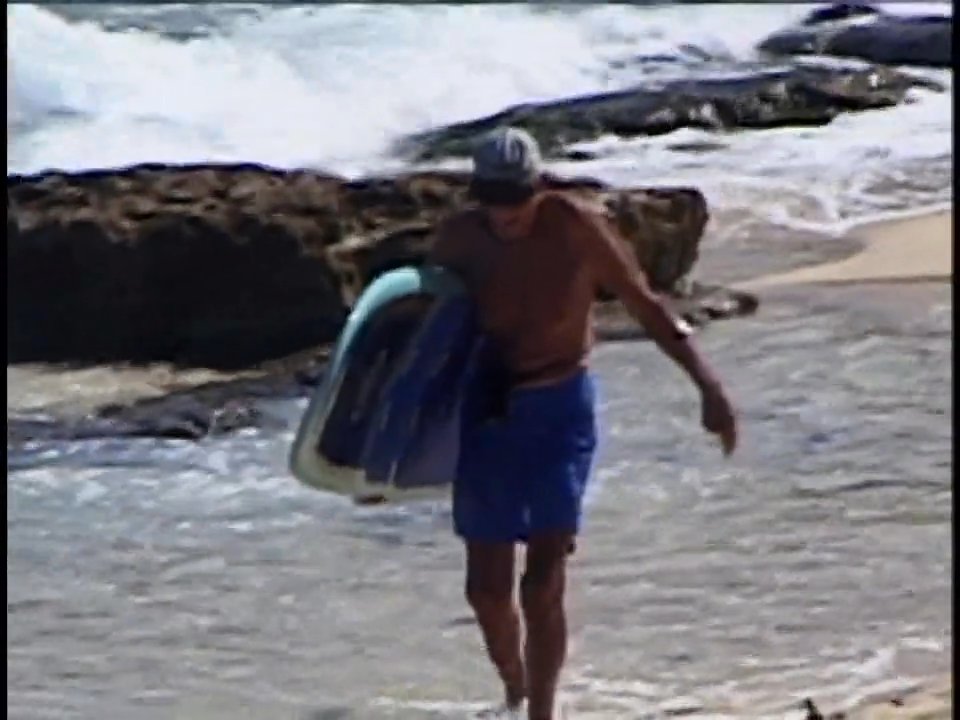 An older man walks from the ocean across the sand while carrying his surfboard, then proceeds along a coastal path through a natural landscape.
