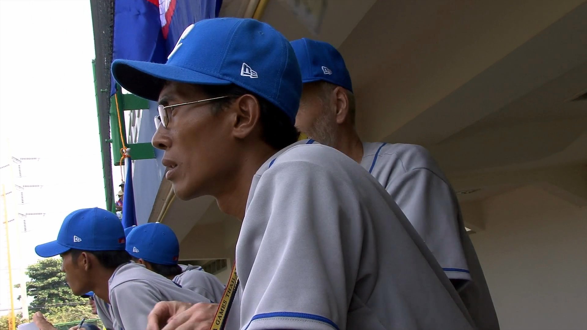 A coach observes a baseball game from the dugout, providing vocal guidance to players while the scoreboard reflects a significant score lead for Indonesia over Cambodia.