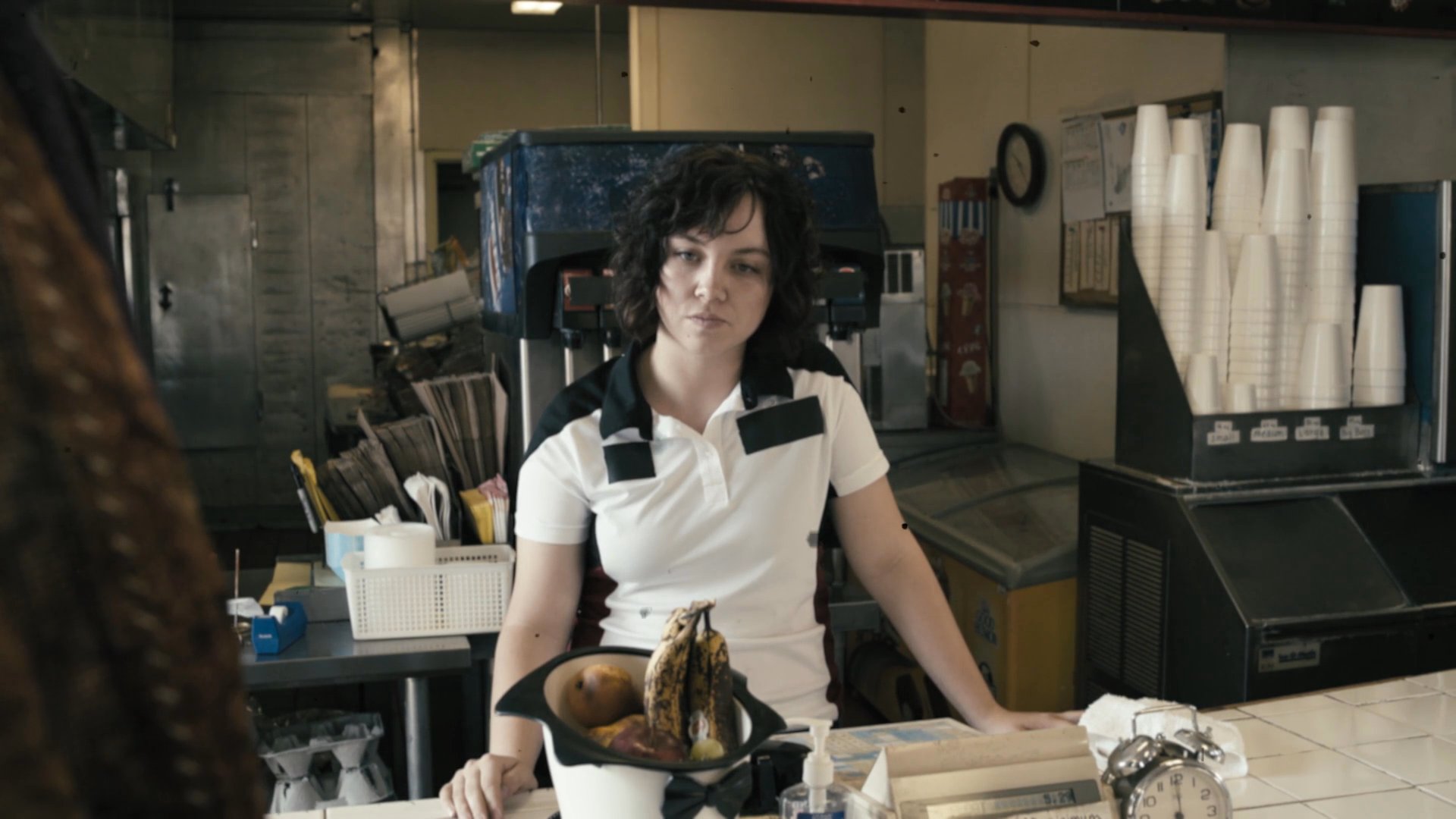 A waitress handles a phone call at a diner counter while a customer draped with a large snake interacts with her. The sequence captures the surreal tension of a mundane setting interrupted by an unusual guest.