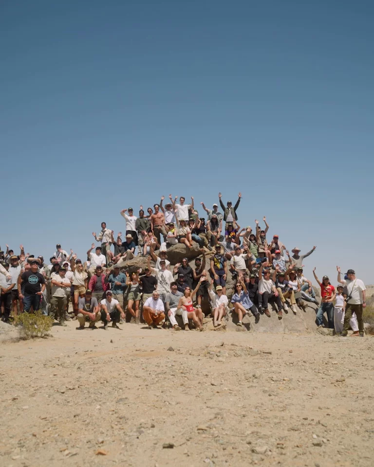 Large group of attendees waving from a giant rock in Joshua Tree