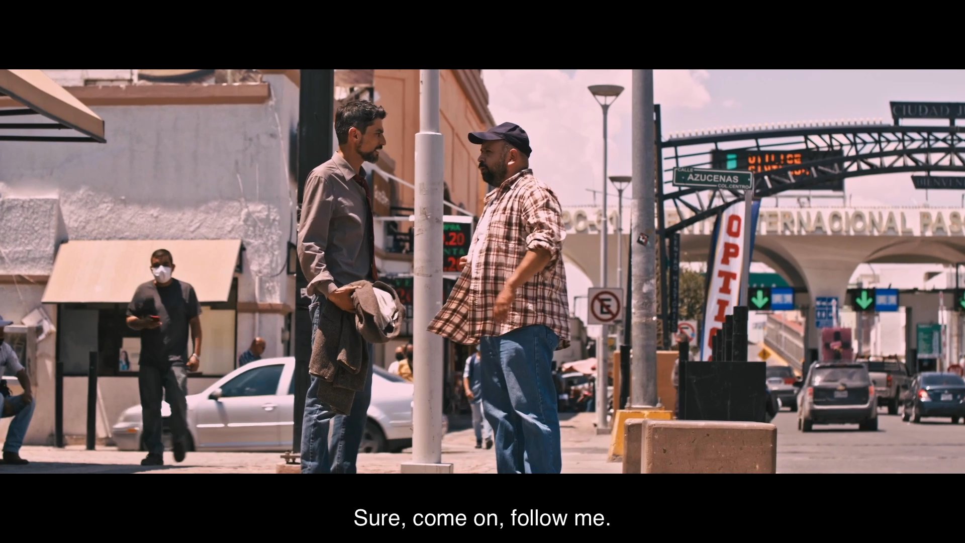 Two men converse at a border crossing checkpoint before navigating through a local street, leading to an encounter with an animated mural.