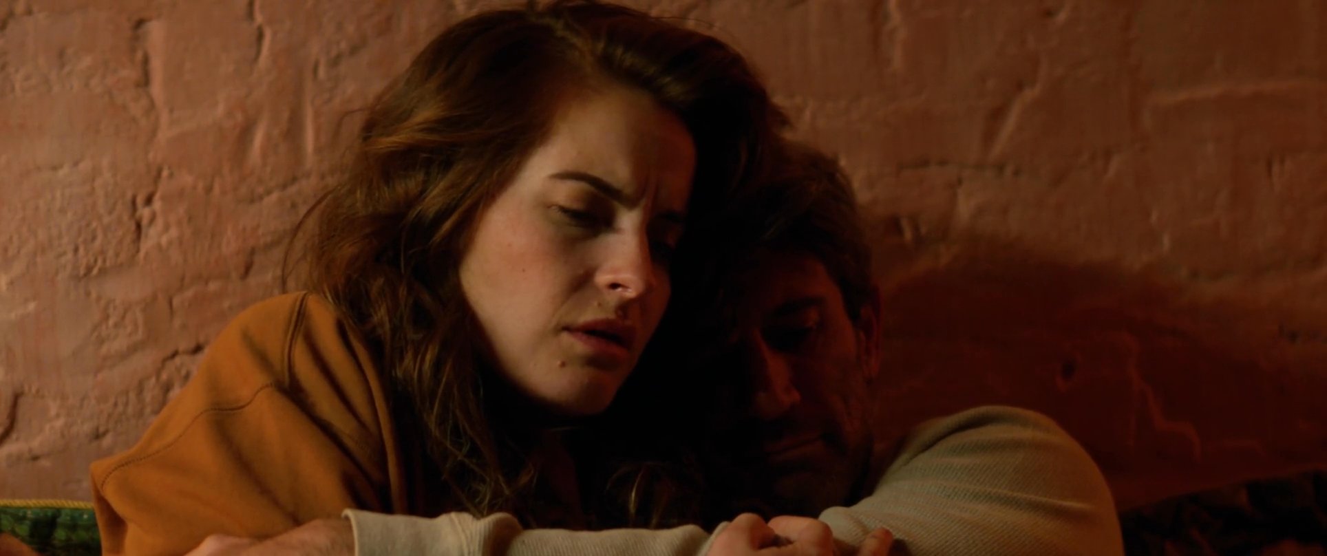 A man offers comfort to a crying woman as they sit together on a bed in a dimly lit room. The sequence captures a tender moment of emotional support and intimacy during a period of personal distress.