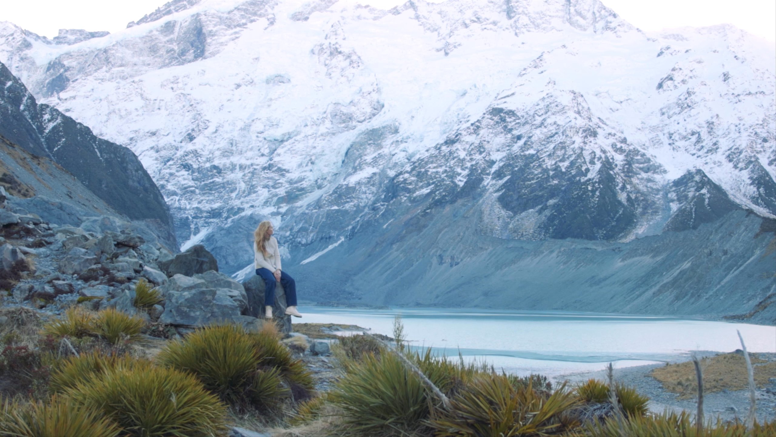 Woman sitting on rock alongside river and snowy mountains in New Zealand