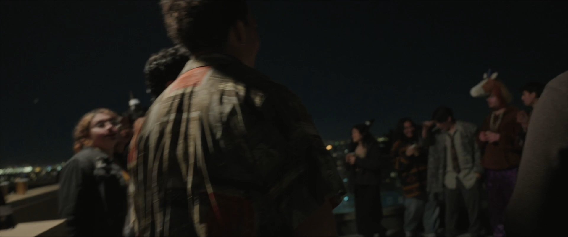 Young adults congregate on a rooftop at night to socialize and celebrate in a lively, casual atmosphere. The group interacts through embraces, conversation, and collective enjoyment of the outdoor setting.