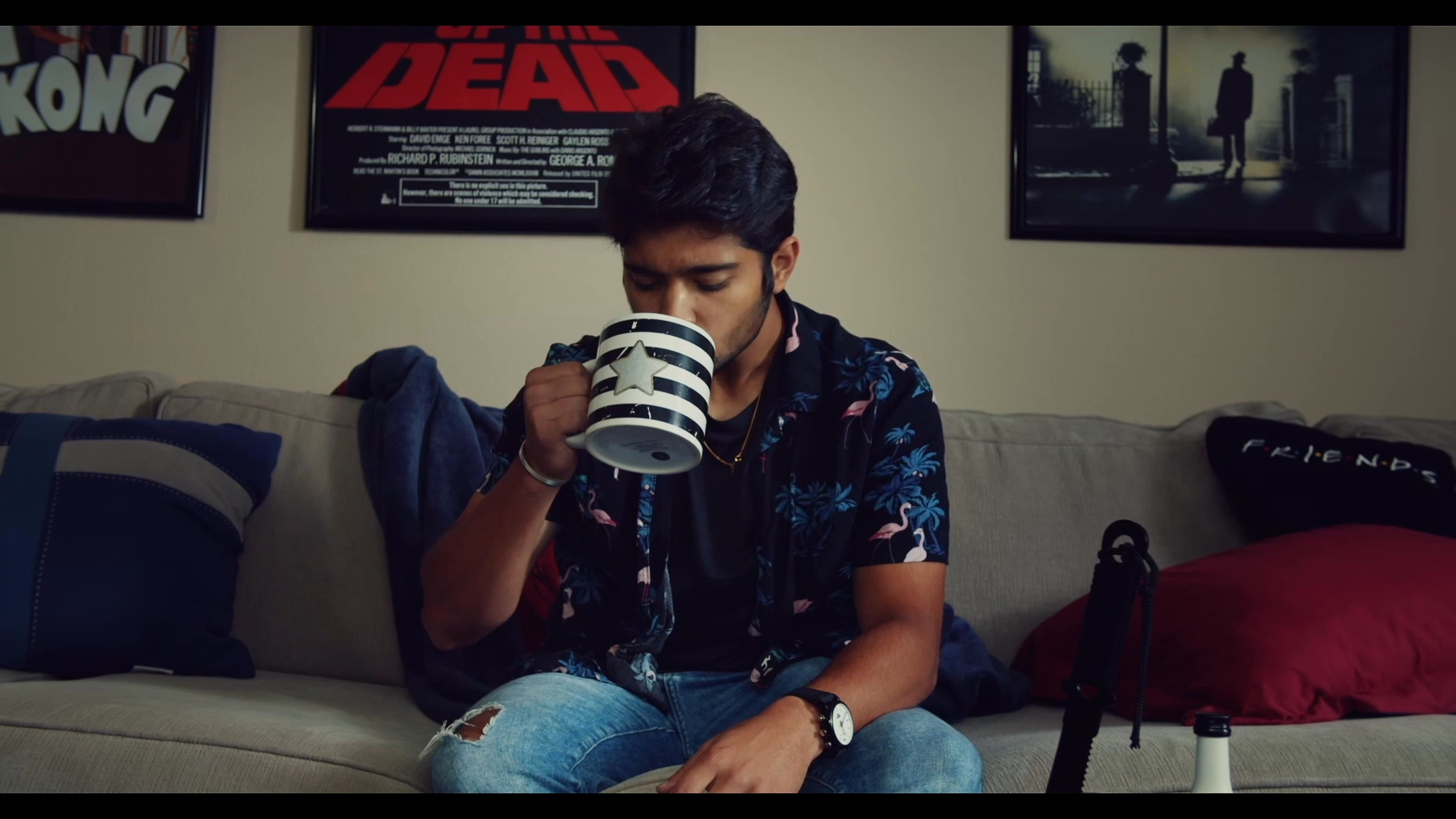 A man sits on a couch drinking coffee while his roommates enter the room, leading to a brief social interaction. The setting transitions from a moment of solitary relaxation to an engaged group dynamic.