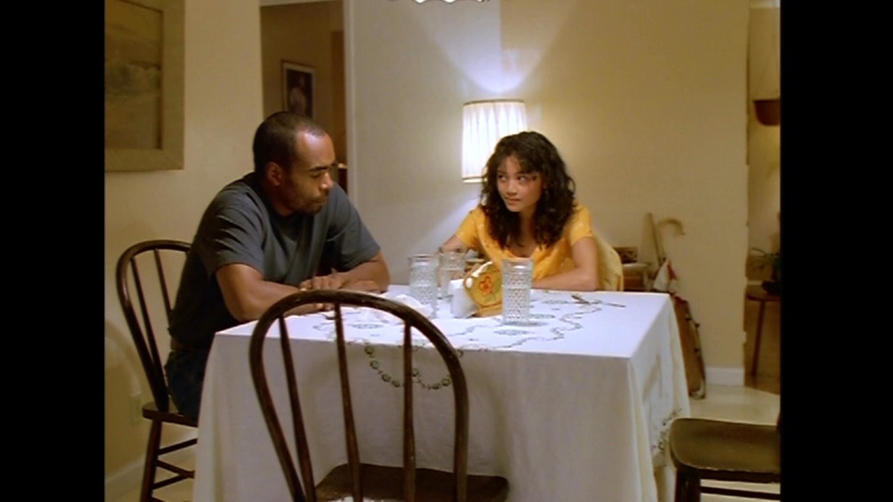 A man and woman sit at a dining table engaged in a serious discussion, while another woman intervenes from the kitchen area. The group later transitions to a living room to continue their exchange.