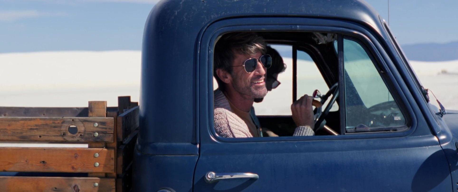 A man drives a vintage blue pickup truck across a vast, white desert landscape and exits the vehicle to survey the surroundings. The scene emphasizes a sense of exploration in a scenic, arid environment.