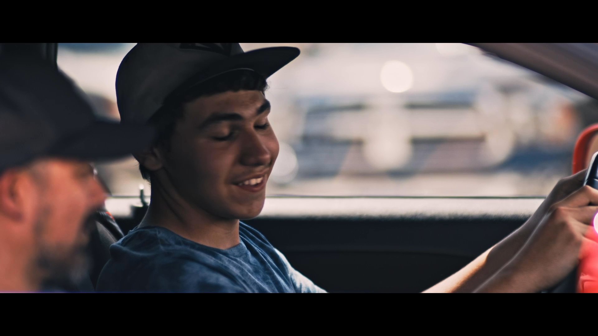 A father and son converse while seated inside a vehicle at a used car dealership in El Paso, Texas. The scene transitions from exterior shots of the dealership lot to an intimate dialogue between the two men.