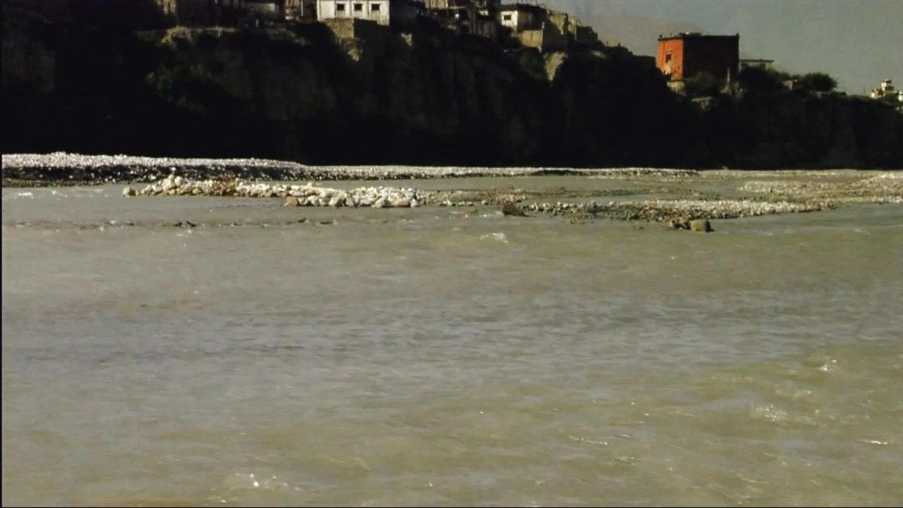 Travelers navigate a remote village and cross a rocky riverbed on horseback against the backdrop of the Himalayan mountain range.