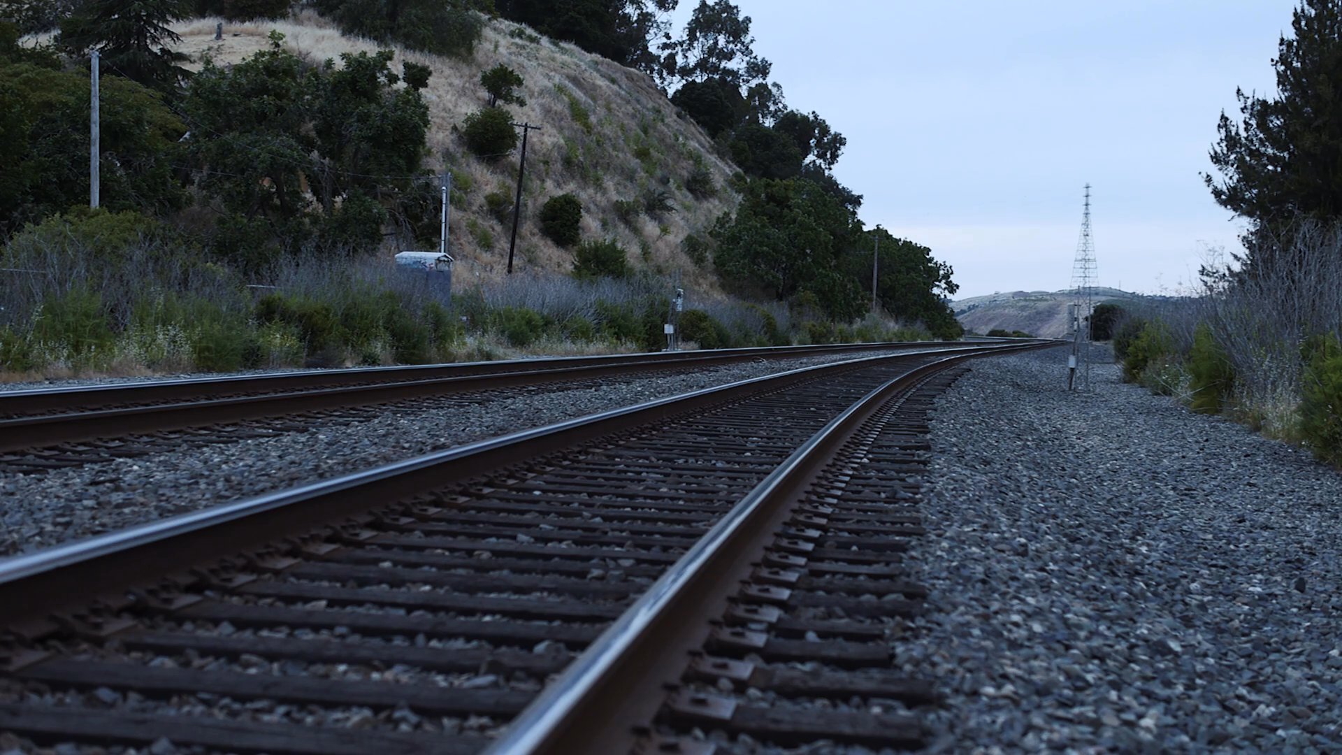 A woman sits contemplatively on railroad tracks near a quiet coastline before returning indoors. The narrative captures a moment of personal reflection set against a somber, natural landscape.