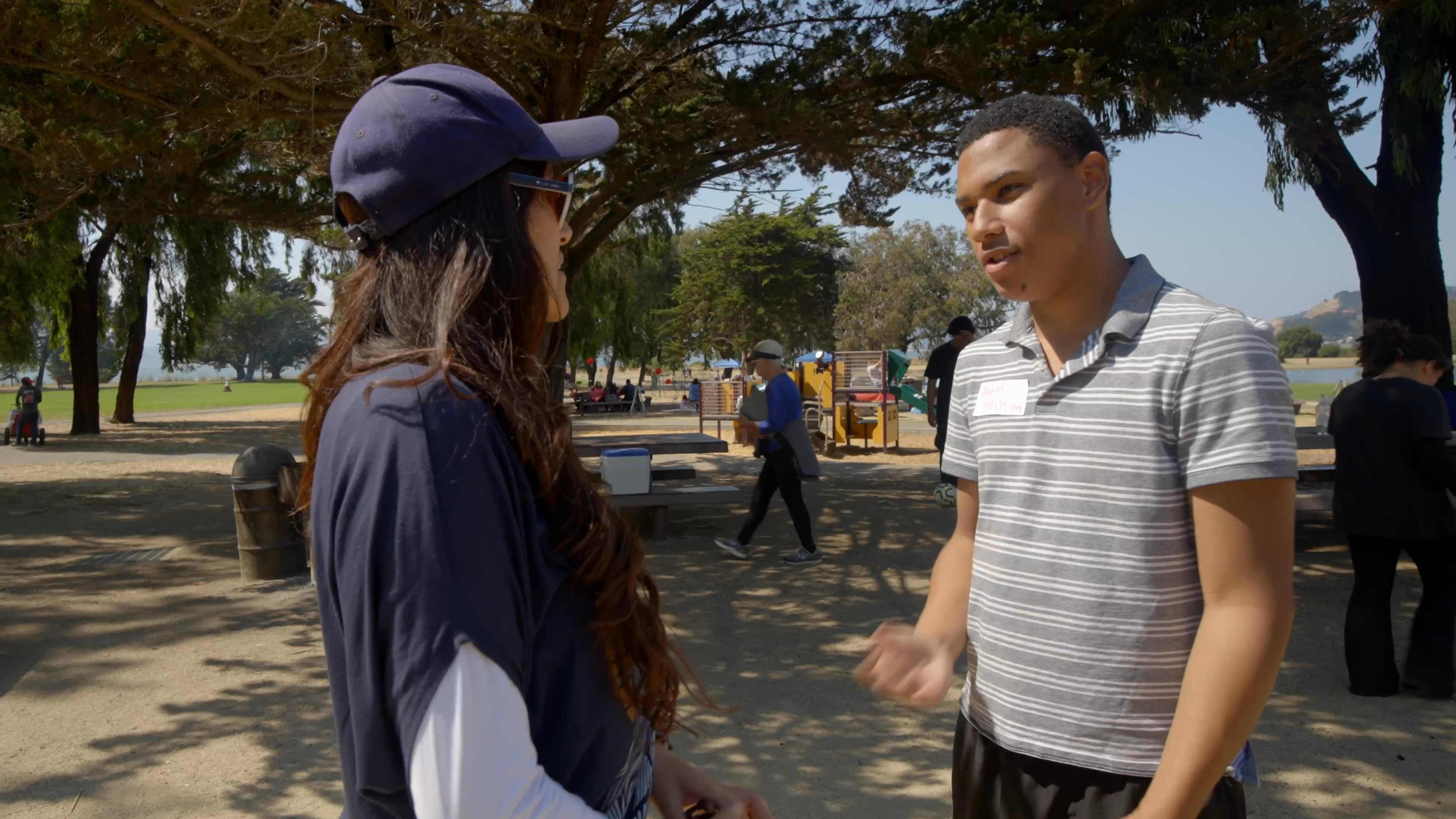 A woman and a man talk beneath the trees in a sunny park, and he gestures as he offers to help. She says she wants others involved with Yes to share an experience she had; the group walks away before she smiles in a lakeside close-up.