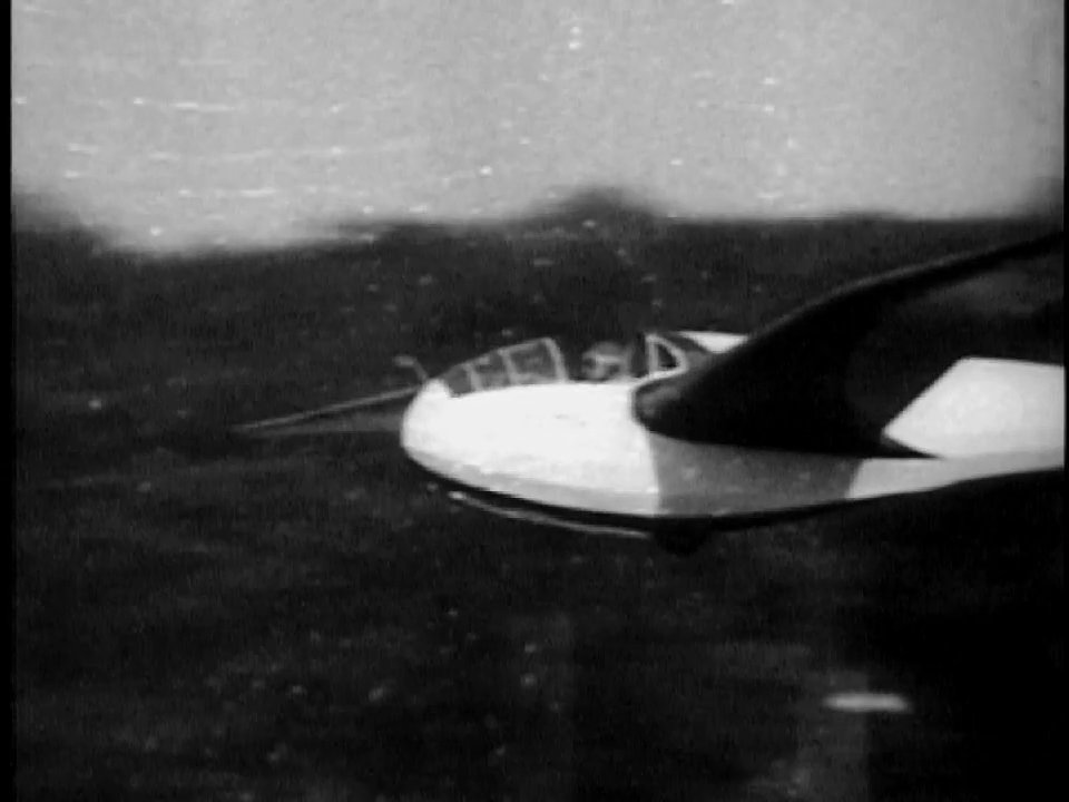 A vintage glider performs a low-altitude flight before a crowd, followed by newspaper headlines announcing the pilot's record-breaking achievement. The sequence documents the successful aviation feat in Wichita, Kansas.