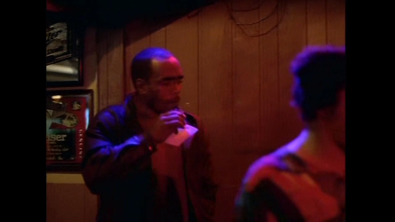 Patrons congregate and socialize within a dim, atmospheric bar setting. Individuals interact, share drinks, and engage in conversation throughout the establishment.