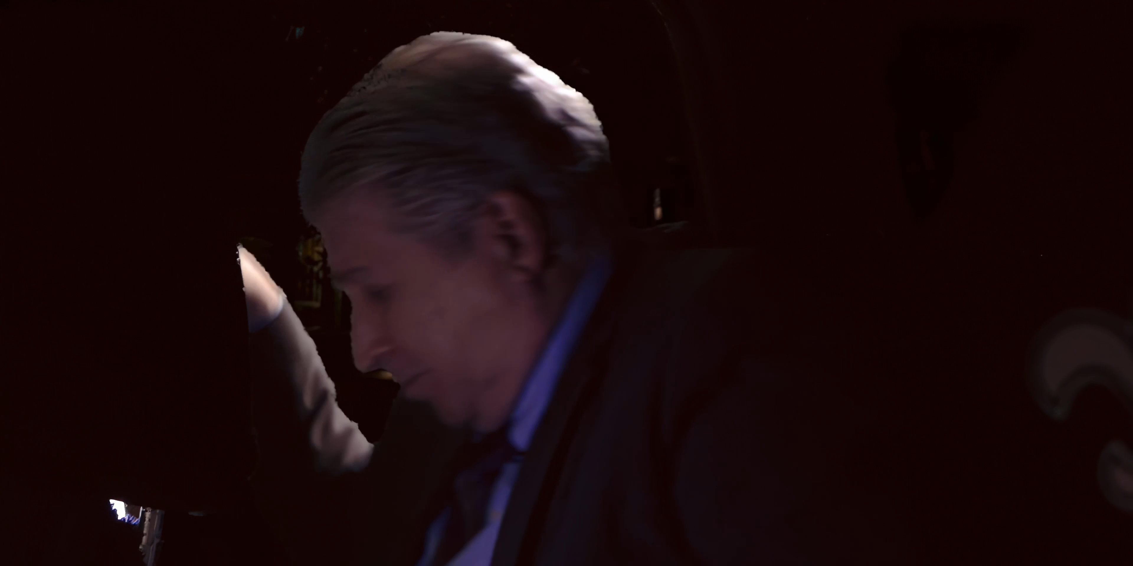 Two men converse inside a car during a night drive, with one wearing a distinct patterned shirt and the other dressed in a business suit.
