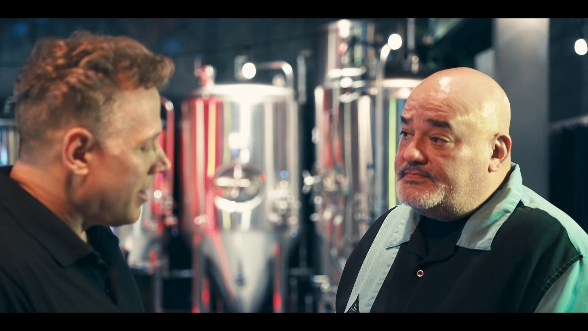 Two individuals engage in a professional discussion within a brewery facility, surrounded by stainless steel fermentation tanks. As the conversation progresses, the setting shifts to include additional staff members, highlighting a collaborative business environment.