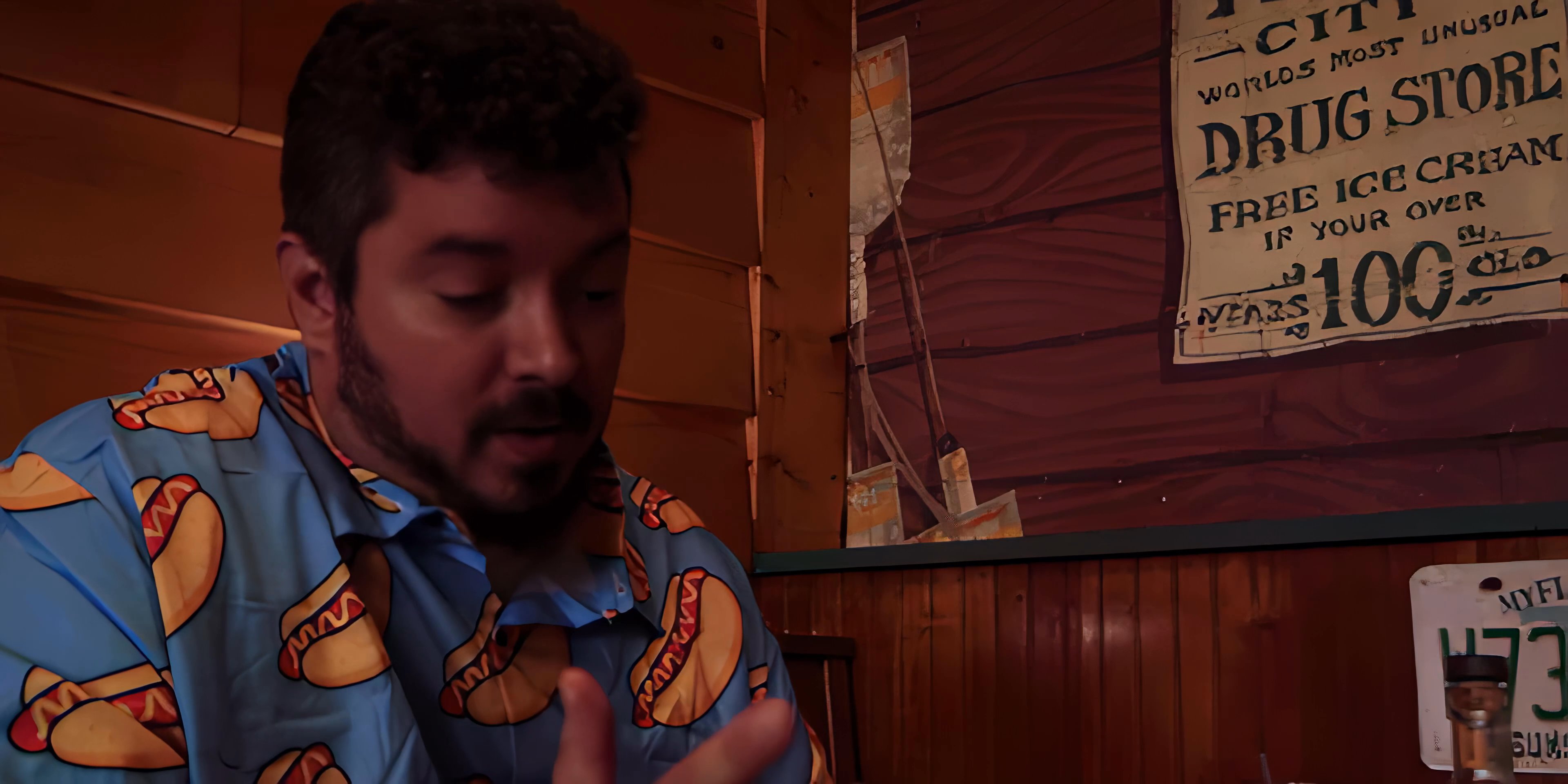 A man in a casual shirt actively gestures and speaks to an off-camera interviewer, engaged in a focused conversation. The setting appears to be a rustic or themed interior with vintage-style wall signage.