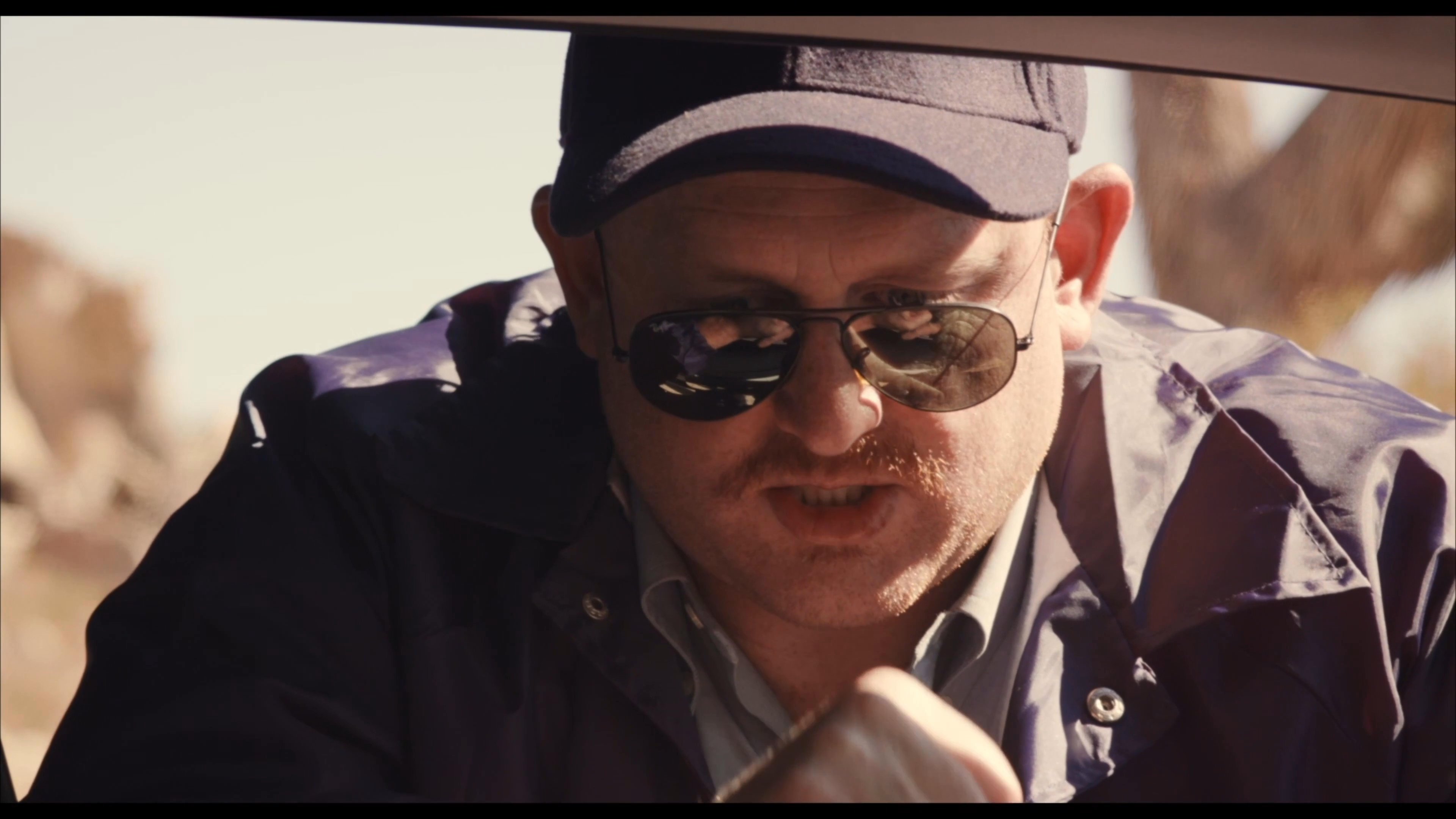 A law enforcement officer approaches a vehicle on a desert highway to question the occupants during a traffic stop. The officer engages in a serious, direct conversation with the driver to assess the situation.