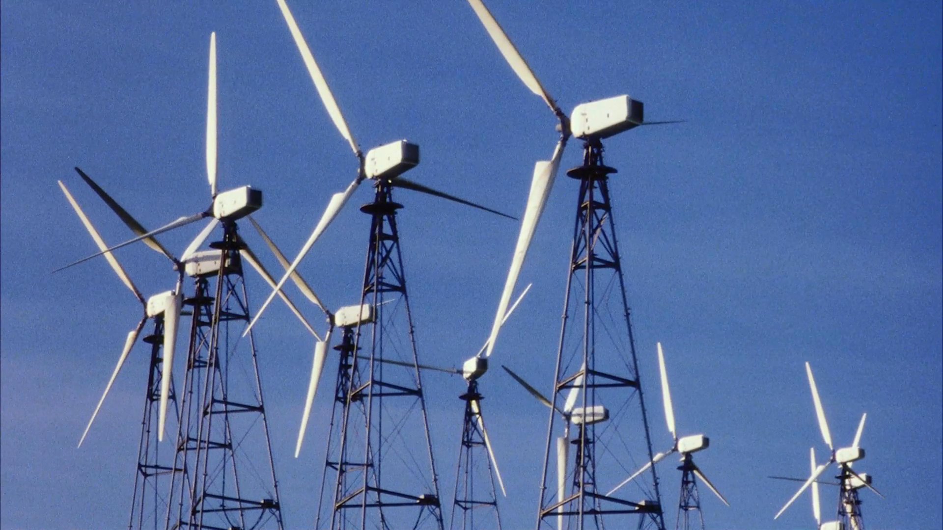 A driver navigates a rural landscape filled with wind turbines before encountering a backpacker waiting on the roadside.