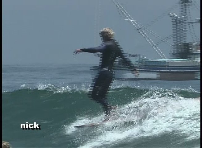 A surfer wearing a wetsuit rides a longboard along a breaking wave with fishing vessels visible in the background. The individual demonstrates balance and control while navigating the swell.