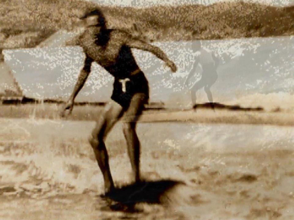 An athlete navigates ocean waves on a surfboard in vintage archival footage. The sequence captures the transition from beach preparation to active surfing performance.