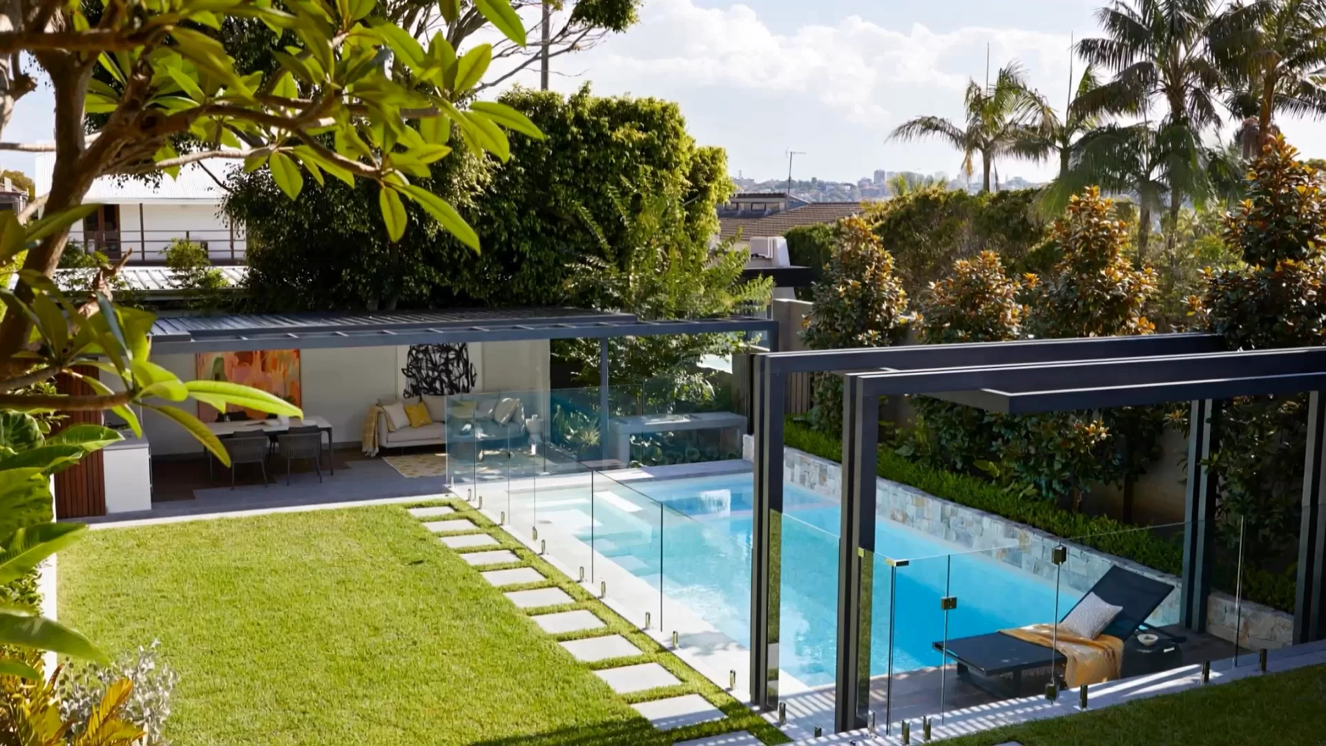 Elevated view over a tropical backyard pool with glass fencing lawn and an open pavilion framed by palms