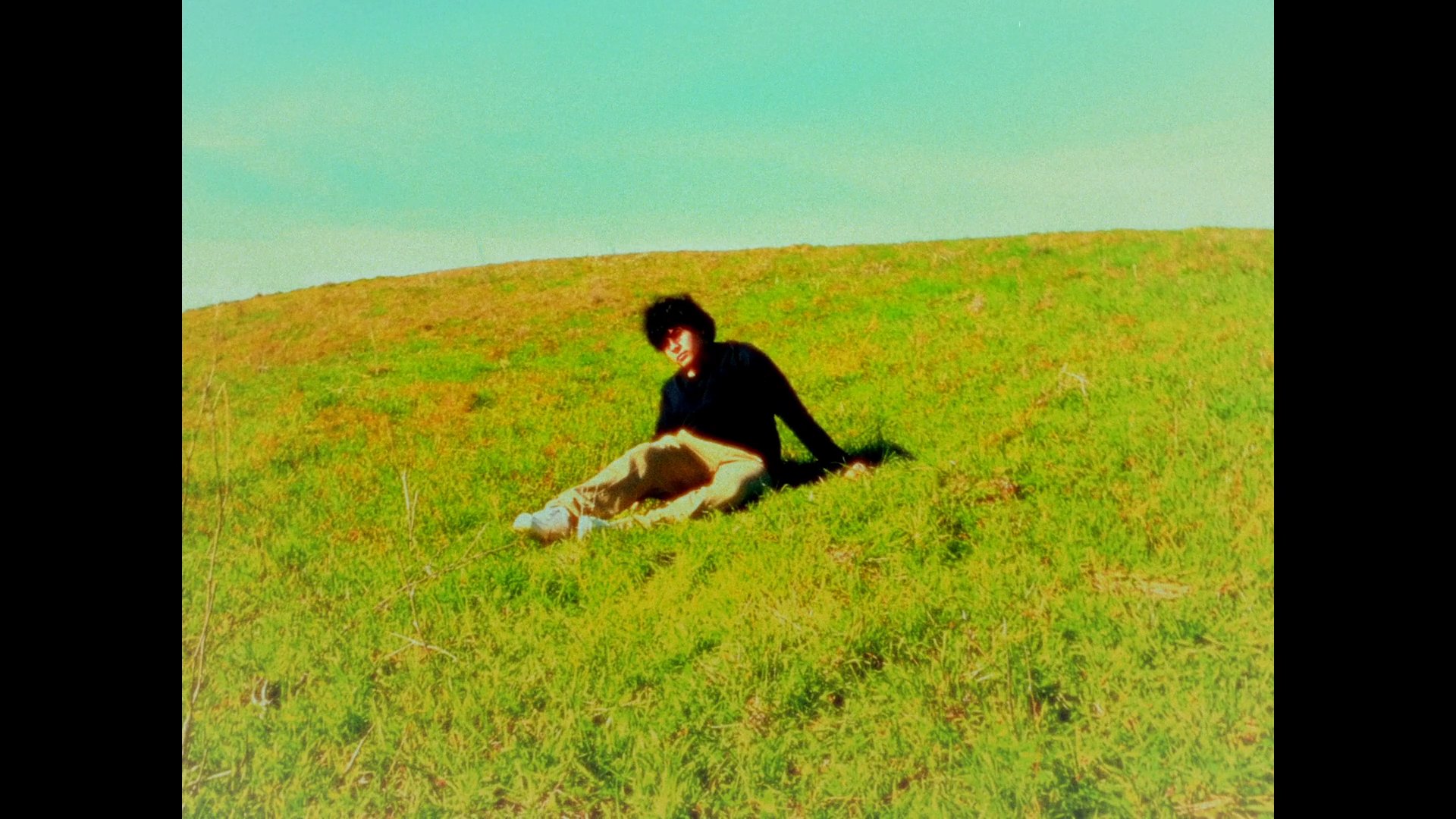 A man sits and rests on a sunlit grassy hill, observing his surroundings in a tranquil outdoor setting.