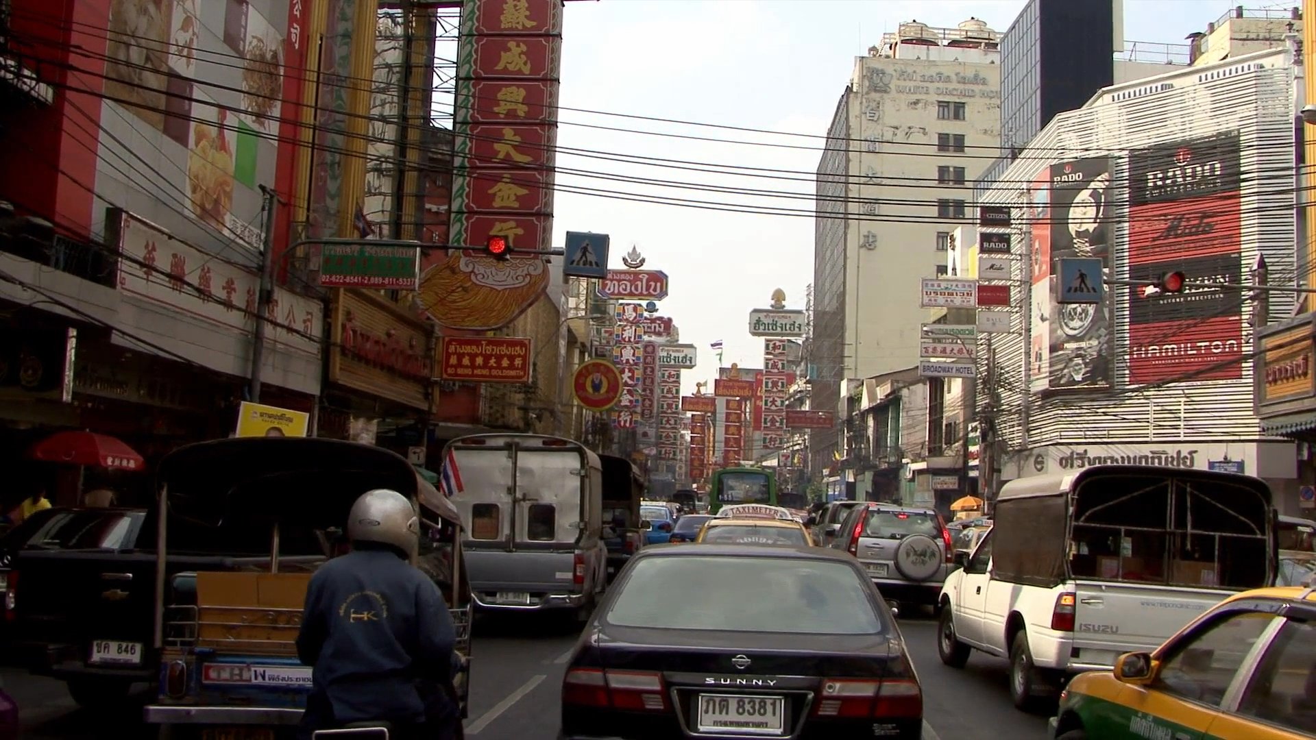A motorcyclist maneuvers through heavy, dense city traffic in the crowded Yaowarat district of Bangkok. Surrounded by cars, buses, and commercial signage, the rider navigates the congested streets during daylight hours.