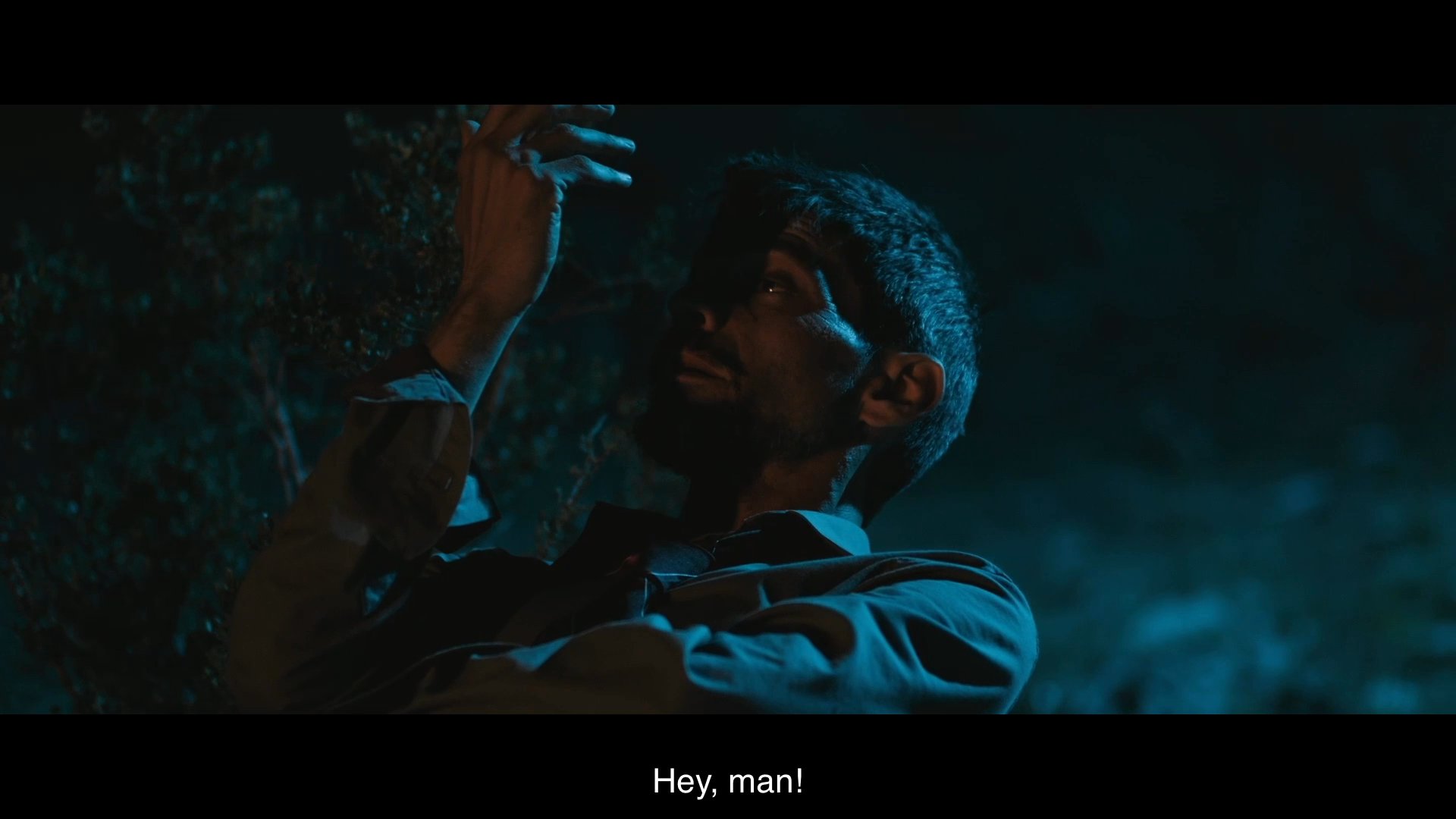 Two individuals encounter one another at a secluded campfire in the wilderness at night, leading to a brief verbal exchange. The sequence captures a tense yet conversational moment between the subjects in a dim, outdoor setting.