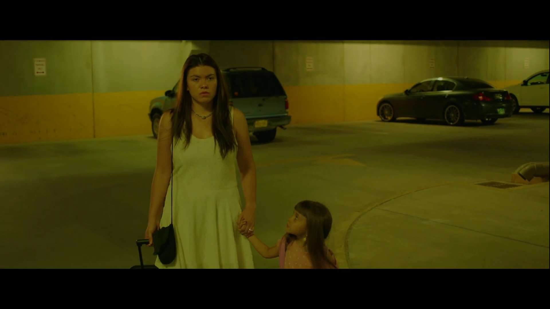 A woman speaks anxiously on a mobile phone while walking with a young child through a dimly lit parking garage at night. They carry luggage, suggesting a departure or urgent transit.