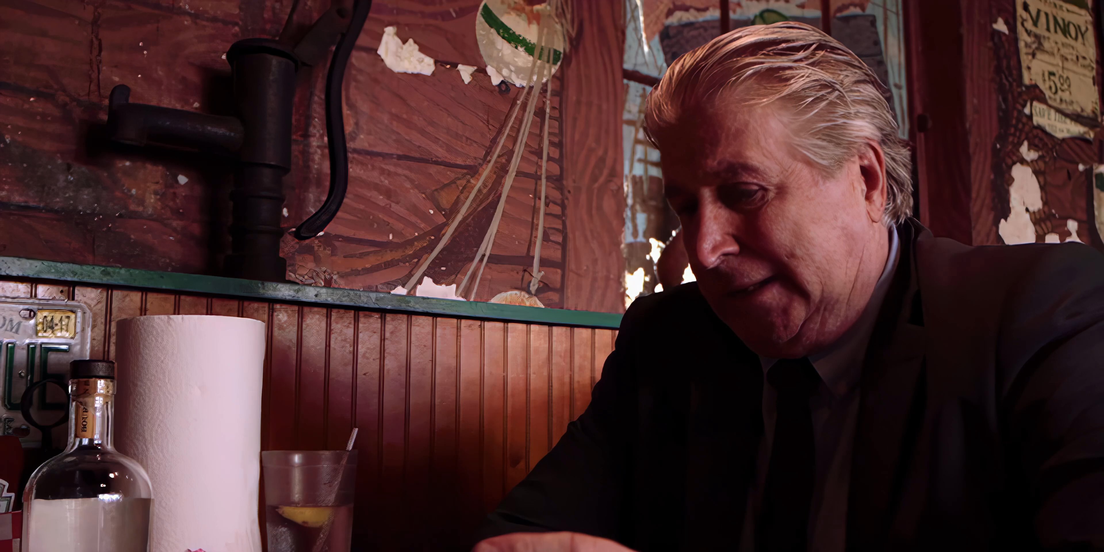 Two men engage in an intense conversation while seated at a restaurant booth. The older man, dressed in a suit, gestures expressively while speaking to the man wearing a patterned shirt.