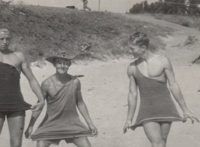 An elderly man shares memories while viewing historical footage of people in period-appropriate swimwear on a sandy beach. The clip juxtaposes his present-day reflection with archival film of a past coastal outing.