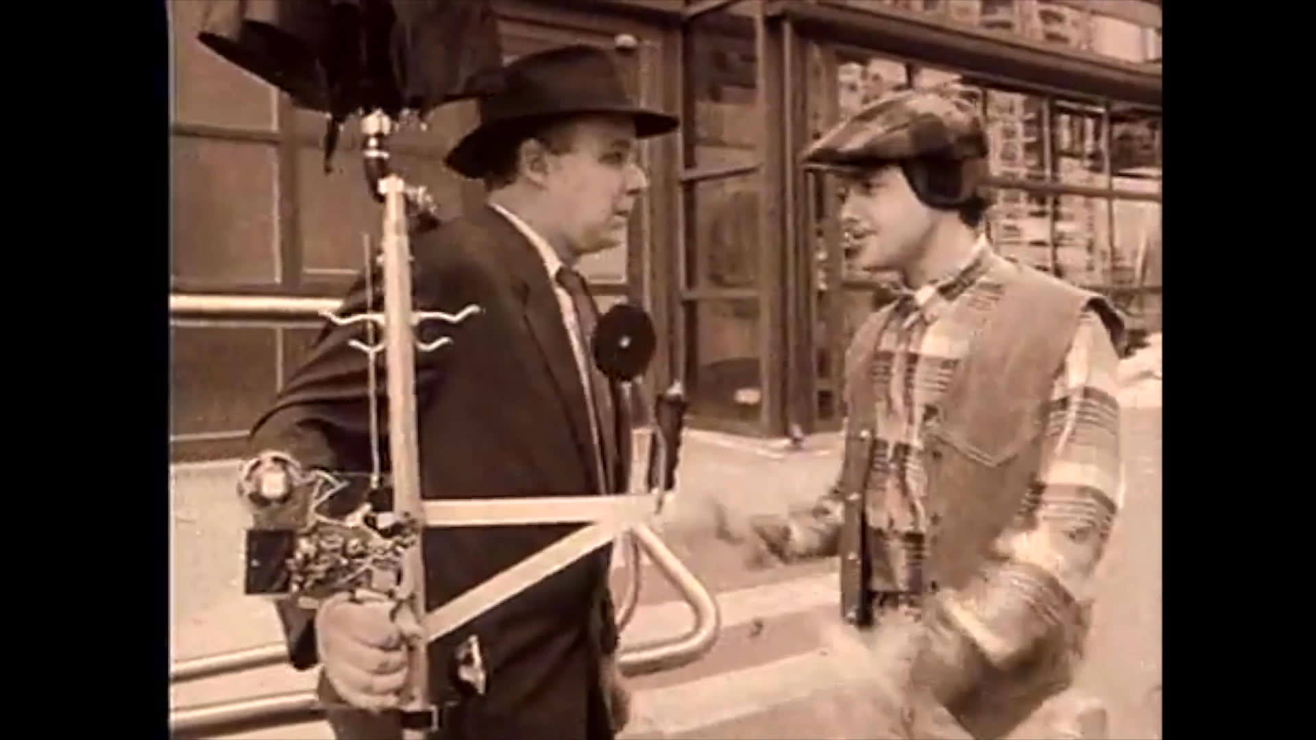 A camera operator in a suit navigates a set carrying specialized equipment while engaging in a professional interaction with a production crew member.