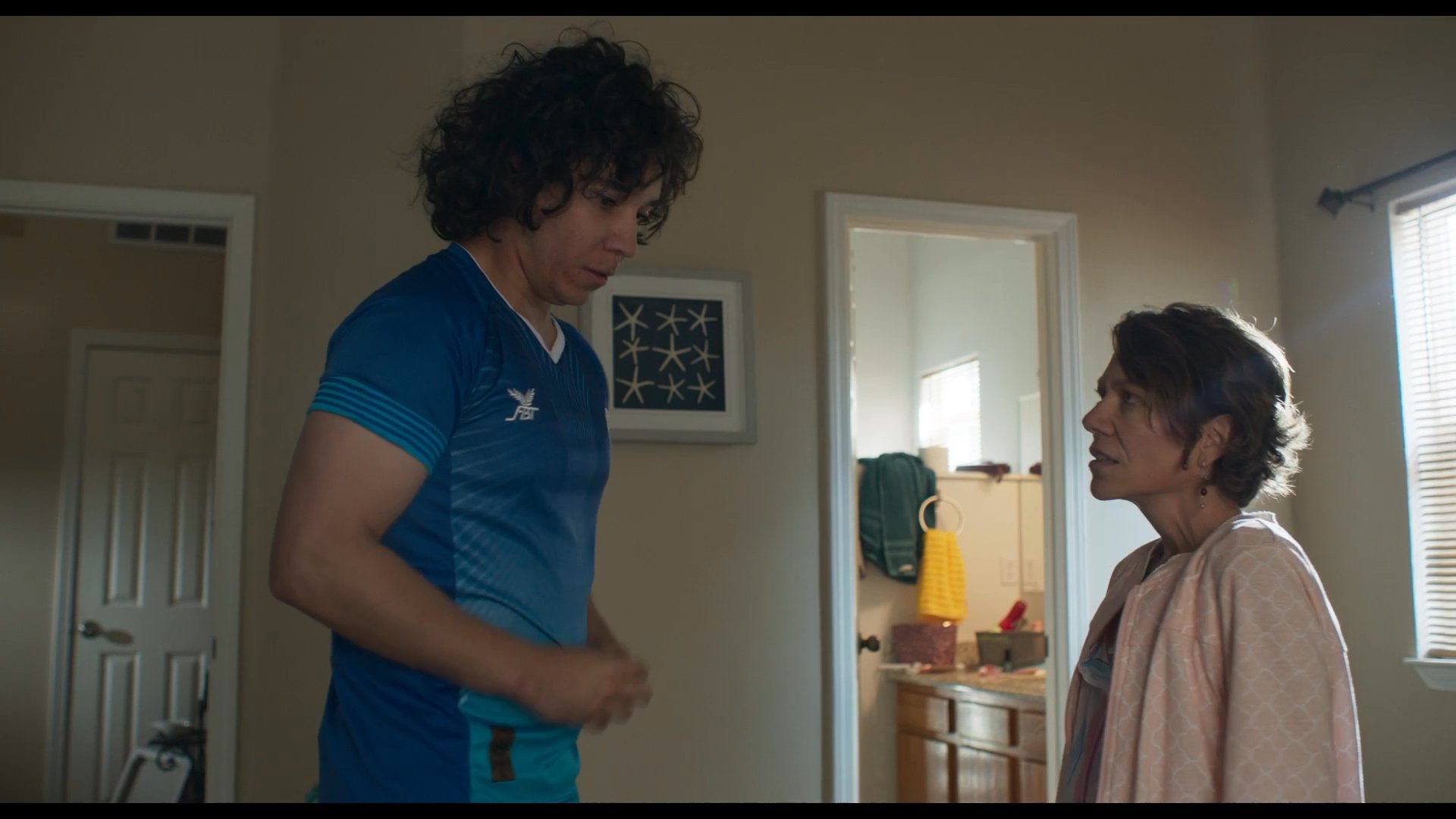 A mother and her son engage in a tender, emotionally charged discussion within a bedroom setting. The interaction transitions from physical comfort to a serious verbal exchange as they sit together on the bed.