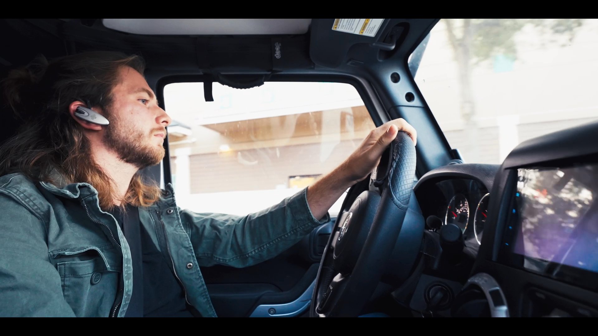 A man with long hair operates a vehicle while wearing a bluetooth headset, occasionally interacting with the car's navigation and entertainment display. He maintains his focus on the road as he travels through an suburban environment.