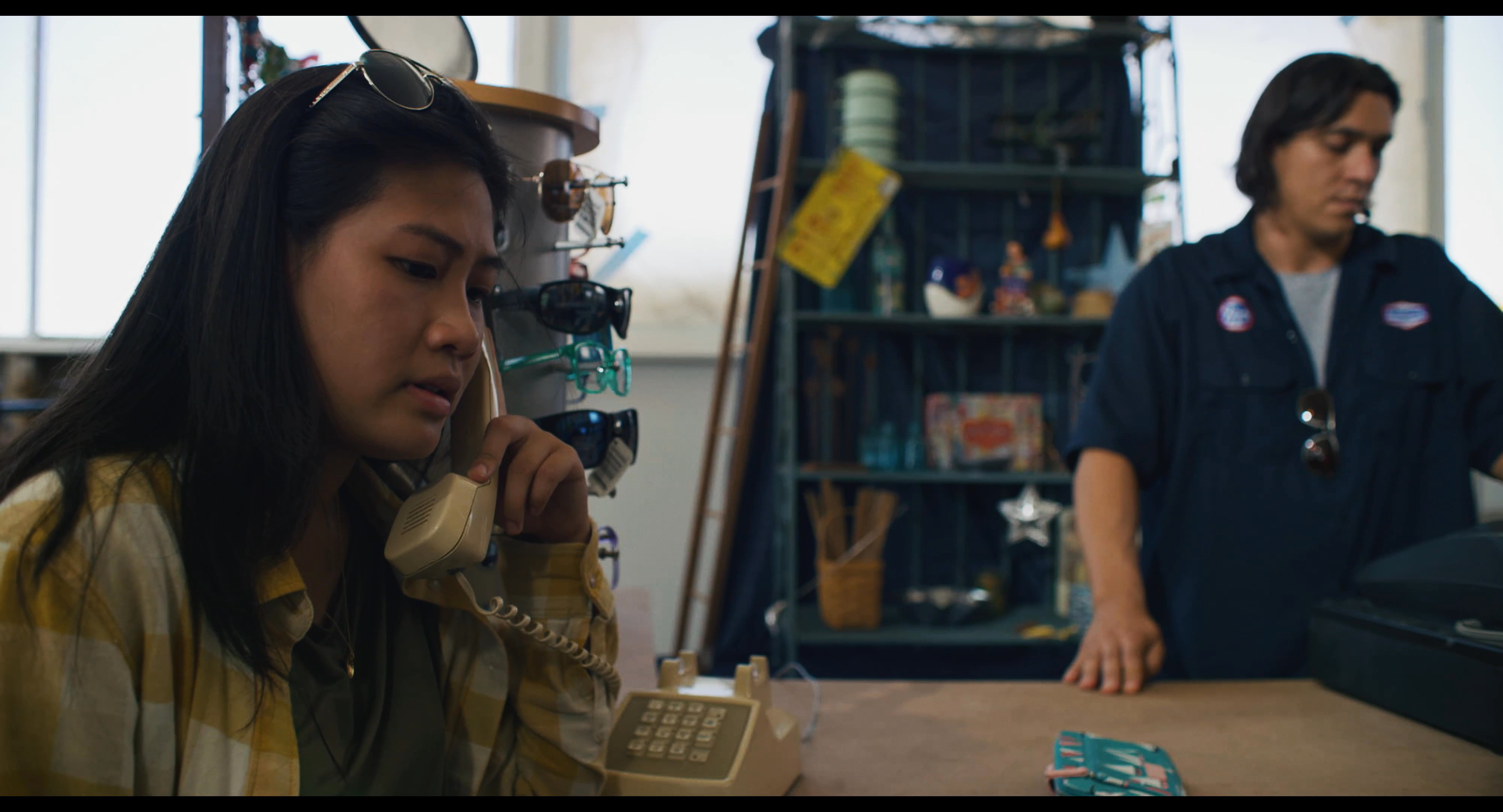 A woman uses a corded landline telephone at a retail counter while a shop employee monitors the transaction. The atmosphere is quiet and focused as the woman conducts her conversation.