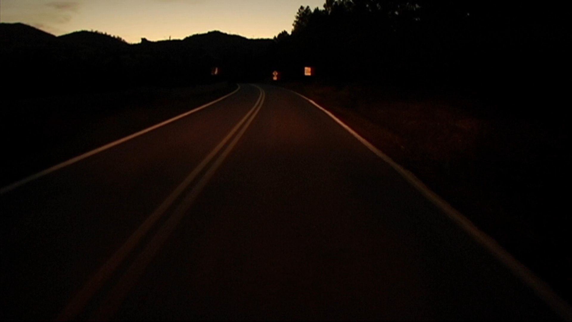 A group of motorcyclists travels along a winding mountain highway at dusk, with their bright headlights illuminating the dark road ahead.