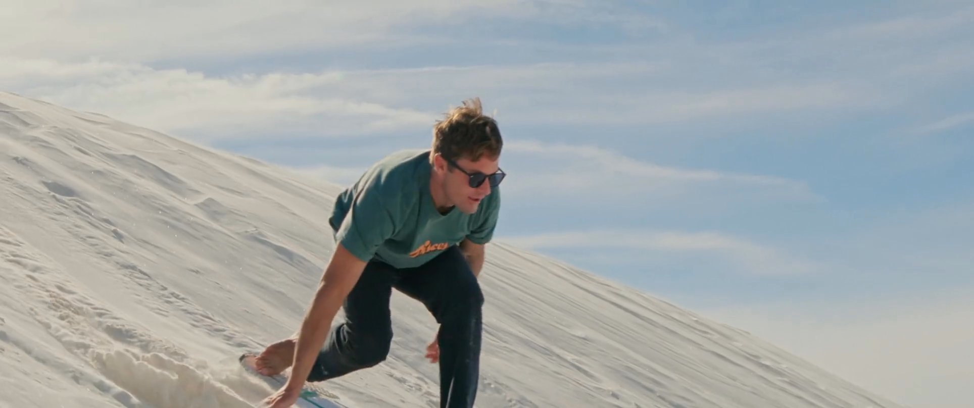 Individuals enjoy recreational sandboarding down the steep, pristine slopes of a white sand dune landscape. The activity captures the dynamic motion and technical balance required for traversing the desert terrain.