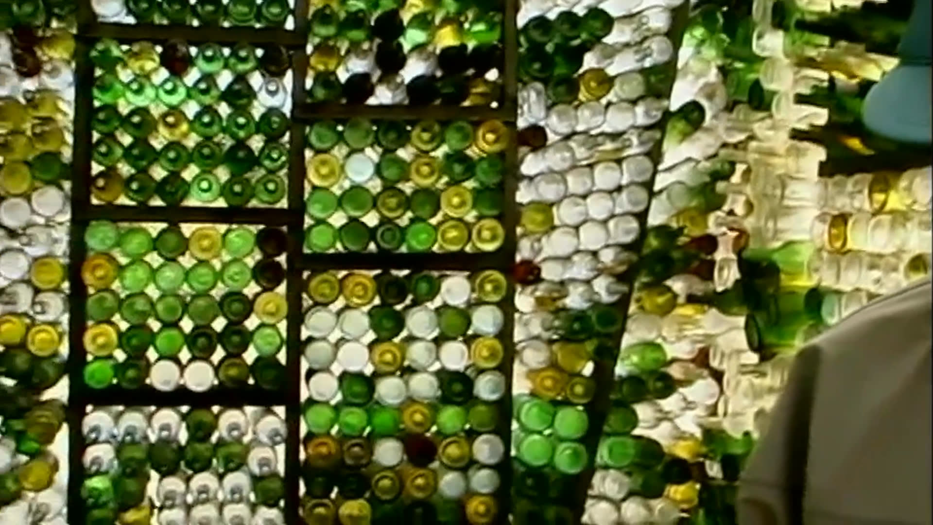 A woman reflects on creating an art installation as an older man stands beside a wall densely arranged with glass bottles. The sequence moves to close views of a large circular sculpture and a small structure set in an open field.