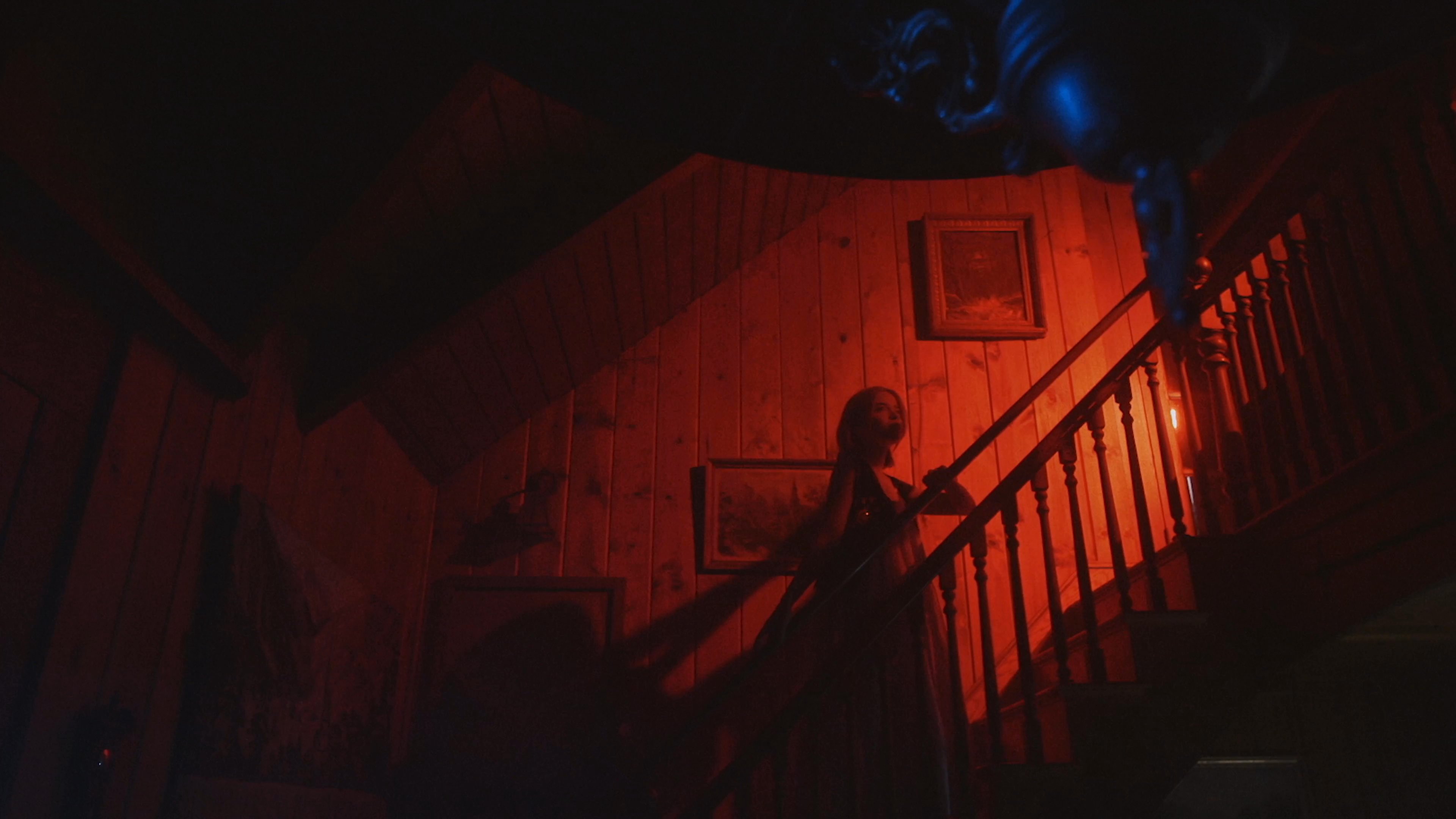 A woman in a white dress traverses a dimly lit, red-hued staircase within a shadowy, atmospheric home. The scene builds tension through stark contrasts of light and dark, evoking a classic gothic horror aesthetic.