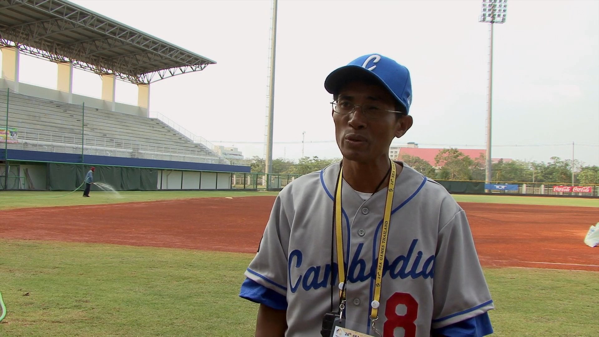 A baseball player wearing a team uniform provides commentary while standing on a professional baseball field. He gestures while discussing team activities against the backdrop of an empty stadium.